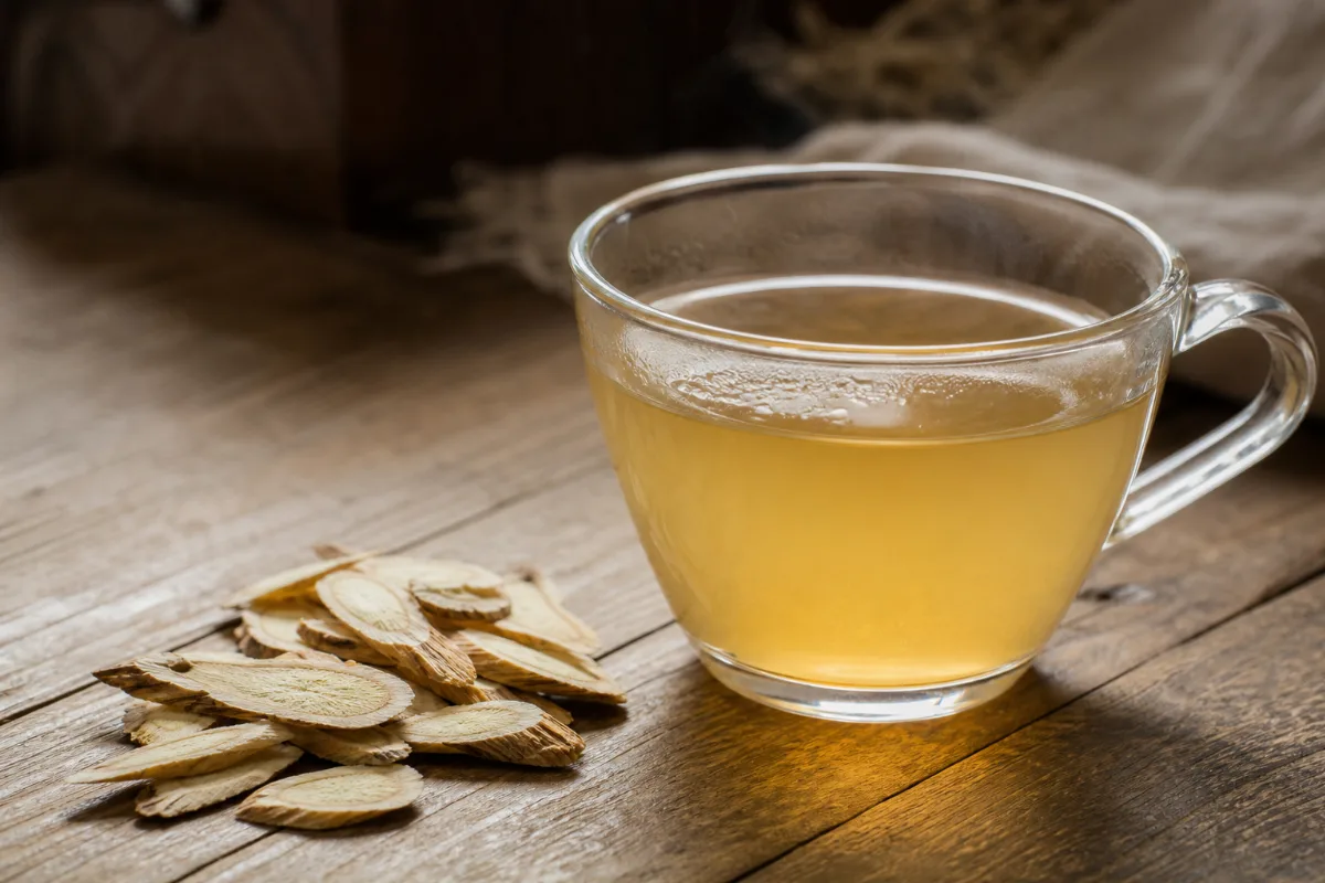 Slices of dried astragalus root beside a cup of pale yellow astragalus tea